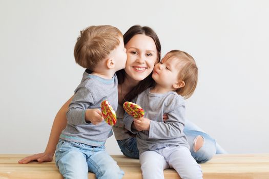 Portrait of a happy mother and her two little children - boy and girl. Happy family against a white background. Little kids kissing mother. Children with lollipops.