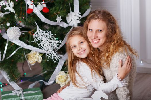 Portrait of happy smiling little girl with mother sitting near Christmas tree.