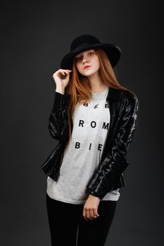 Portrait of beautiful young girl wearing black felt hat and leather jacket in front of a dark gray background