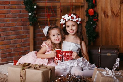 Little girls with Christmas knitted cups. Little girls in hair accessories sitting on floor in beautiful Christmas decorations. New year celebration. Happy girls and family.