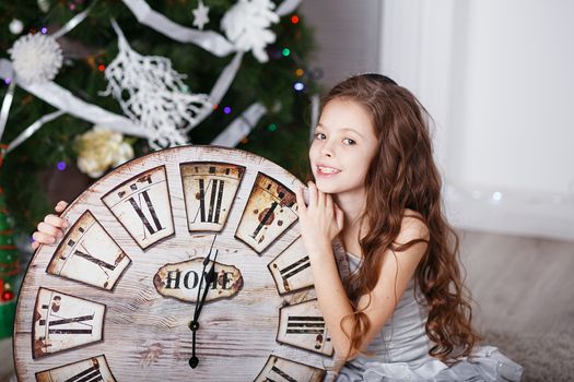 Portrait of Beautiful little girl with long curly hair in silver dress with a big clock waiting for the new year. New year preparation