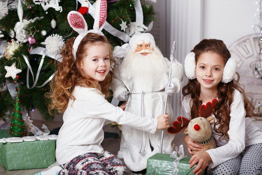 Little girls in comfortable home clothes sitting on floor in beautiful Christmas decorations. Two little sisters decorating Christmas tree with fir-cone. New year preparation. Happy girls and family.