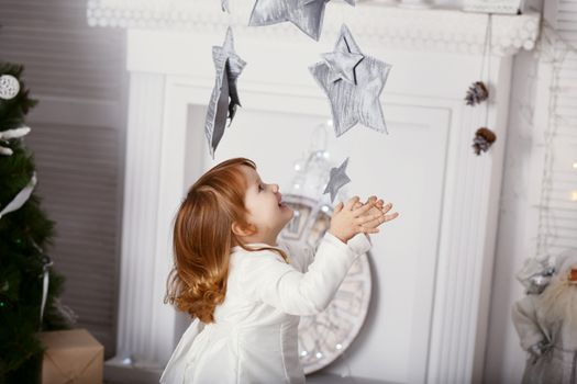 Portrait of a beautiful little girl in a white dress in the interior with Christmas decorations. Baby girl grabs and catches toy star - an element of a Christmas decor