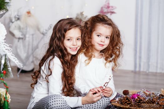Little girls in comfortable home clothes sitting on floor in beautiful Christmas decorations. Two little sisters decorating Christmas tree with fir-cone. New year preparation. Happy girls and family.