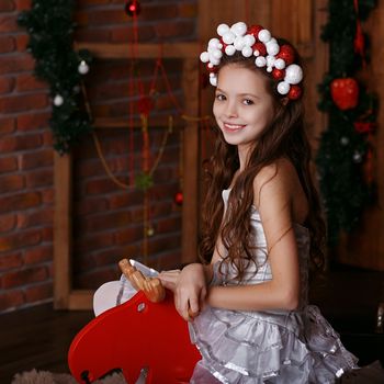 Portrait of a lovely little girl in hair band. Portrait of cute little girl among Christmas decorations. Girl riding a rocking deer. Rocking deer chair for kids ride playing