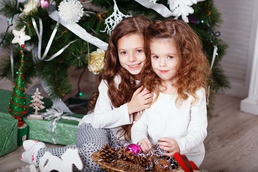 Little girls in comfortable home clothes sitting on floor in beautiful Christmas decorations. Two little sisters decorating Christmas tree with fir-cone. New year preparation. Happy girls and family.