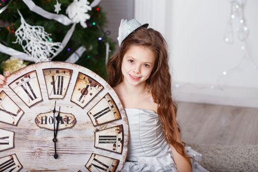 Portrait of Beautiful little girl with long curly hair in silver dress with a big clock waiting for the new year. New year preparation