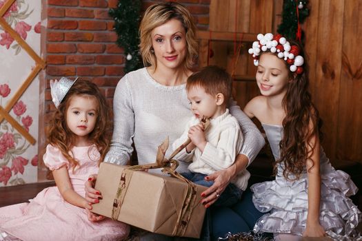 Mother and her two little daughters and son opening a Christmas gift. Family on Christmas eve. Mother and little kids opening Xmas presents. Children with gift boxes. Cozy winter evening at home.