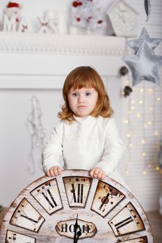 Portrait of a beautiful little girl in a white dress in the interior with Christmas decorations. Baby girl with big clock dial