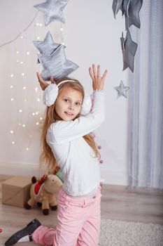 Portrait of a beautiful little girl in a in earmuffs in the interior with Christmas decorations. Girl grabs and catches toy star - an element of a Christmas decor