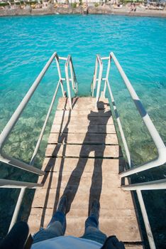 Old vintage metal wooden stairs leading to blue turquoise sea water from beach. Beautiful summer day. Swimming pool with grunge retro stairs on the ocean coast. Top POV, Point of view shot.