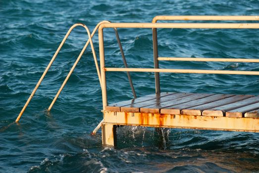 Old vintage metal wooden stairs leading to blue turquoise sea water from beach. Beautiful summer day. Swimming pool with grunge retro stairs on the ocean coast. Side View.