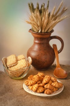 Small cookies on a plate and burlap. Cookie jar, wooden spoon,jug with ears of rye and wheat in the background, warm light and bokeh. A gradient background.