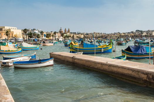 Traditional fishing village Marsaxlokk at the island Malta. Many colorful wooden boats in the bay. Church in the background.