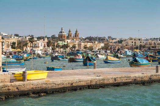 Traditional fishing village Marsaxlokk at the island Malta. Many colorful wooden boats in the bay. Church in the background.