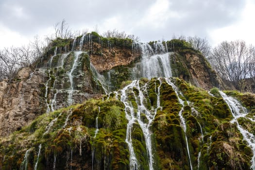 View of a waterfall, flowing from high inside meadow area, with green trees and plants around.