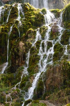 View of a waterfall, flowing from high inside meadow area, with green trees and plants around.