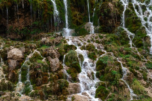 View of a waterfall, flowing from high inside meadow area, with green trees and plants around.