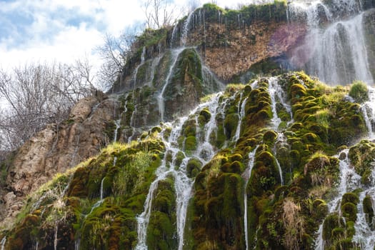 View of a waterfall, flowing from high inside meadow area, with green trees and plants around.