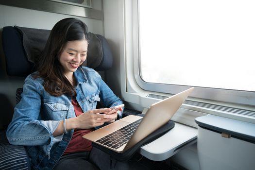 Asian woman smiling at smartphone with laptop on train, copy space on window