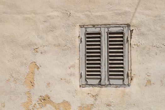 Old wall with a peeled off facade and a small window with a wooden closed shutter. A shadow of a cable crossing the window.