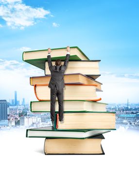 Businessman hanging on top of stack books city background.