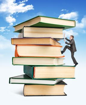 Pile of books with businessman climbing it on blue sky background