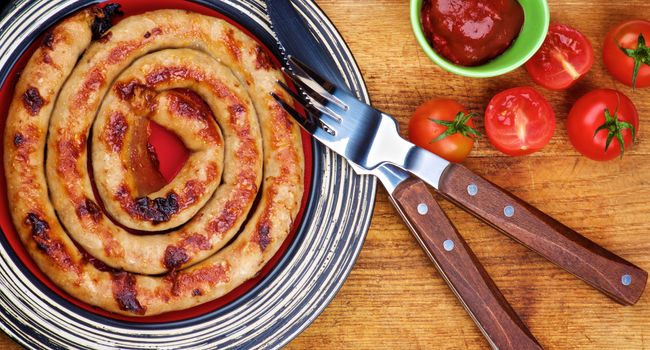 Delicious Grilled Spiral Sausage on Striped Plate with Ketchup, Cherry Tomatoes  and Rustic Fork and Knife on Wooden Cutting Board closeup. Top View