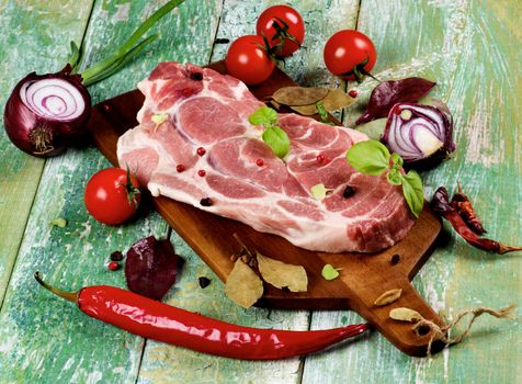Perfect Raw Pork Neck on Wooden Cutting Board with Arrangement of Spices, Herbs, Tomatoes, Red Onion and Fresh Chili Pepper closeup on Cracked Wooden background
