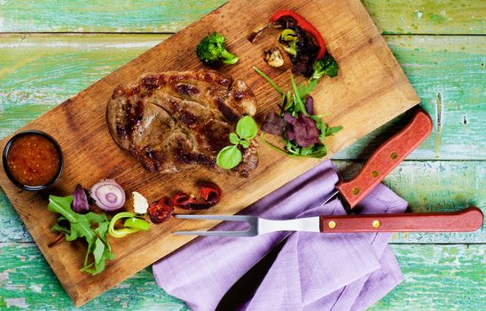 Delicious Roasted Pork Neck with Grilled Vegetables, Fresh Greens and Hot Sauce closeup on Wooden Cutting Board with Meat Fork and Knife. Top View