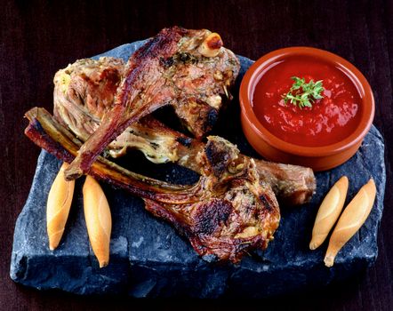 Arrangement of Three Delicious Roasted Lamb Ribs with Bread Sticks and Tomato Sauce on Stone Plate closeup on Dark Wooden background