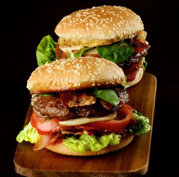Two Tasty Hamburgers with Beef, Bacon, Lettuce, Tomatoes, Basil, Roasted Onion and Juicy Sauce on Sesame Buns on Wooden Cutting Board closeup on Dark Wooden background