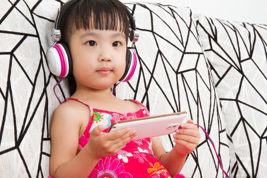 Chinese little girl on headphones holding mobile phone sitting on sofa