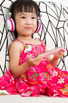 Chinese little girl on headphones holding mobile phone sitting on sofa