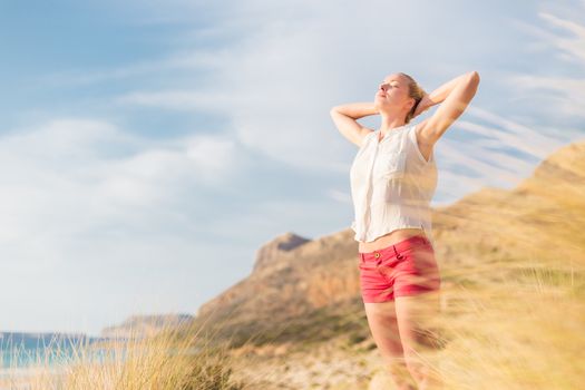 Relaxed woman, arms rised, enjoying sun, freedom and life an a beautiful beach. Young lady feeling free, relaxed and happy. Concept of vacations, freedom, happiness, enjoyment and well being.