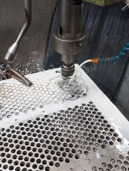 Column drilling machine making holes in a steel plate while being cooled down by cooling liquid. Drill parts blurred due to motion.