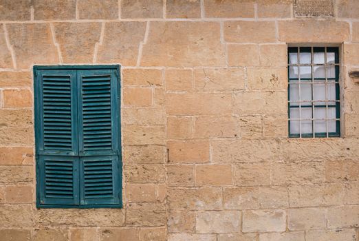 Two windows on the stone wall of an old house in the former capital of island Malta - Mdina. Closed green shutter, the other window without shutter.