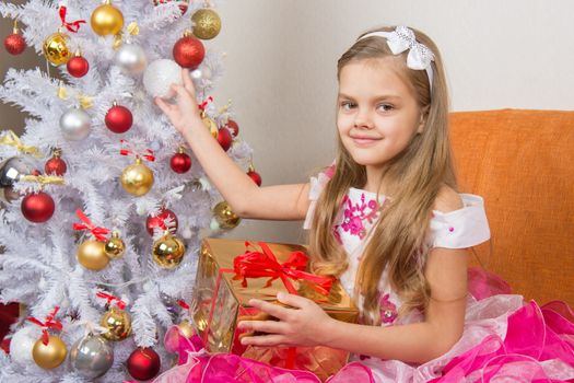 Seven-year girl in a beautiful dress sits with a gift and holding a Christmas ball in hands