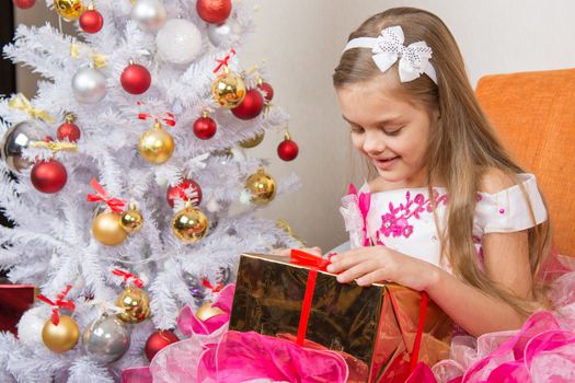 Seven-year girl in a beautiful dress is considering a gift sitting on the couch at Christmas tree
