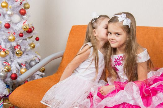 Two girls in beautiful dresses whispering sitting on couch at Christmas tree