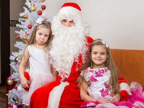 Two girls in beautiful dresses hug Santa Claus sitting on the couch, one of them a little displeased