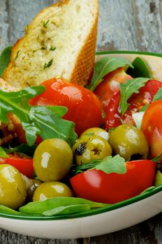 Fresh Tasty Tomatoes Salad with Arugula, Olives and Greens with Garlic Bread in Bowl closeup on Rustic Wooden background