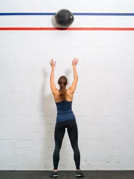 Photo of a young fit woman exercising by throwing a medicine ball up against a wall in a gym.