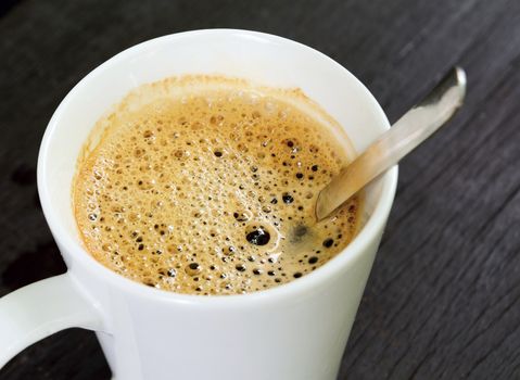 Top view to the white cup with brown coffee and spoon on the brown wooden table.