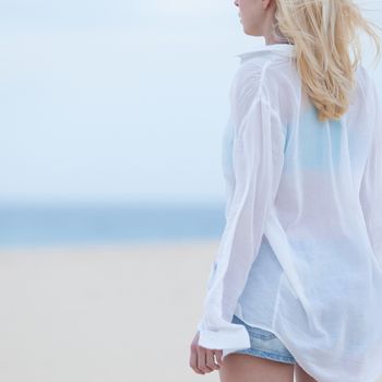 Sensual blonde woman wearing white loose casual shirt on vacations looking down sandy beach in dusk.