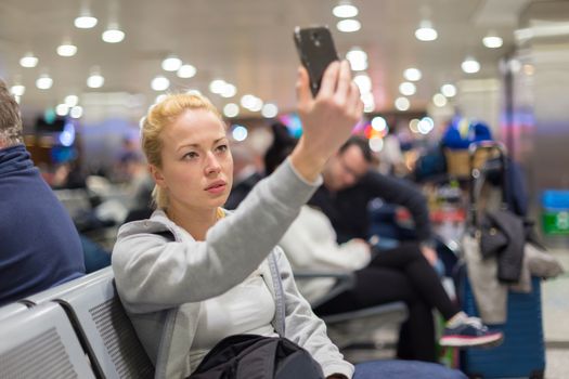 Casual blond young woman taking selfie photo using her cell phone while waiting to board a plane at the departure gates.