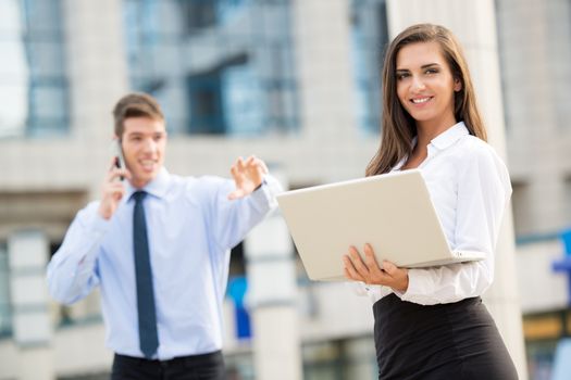 Young business couple standing in front of the building. Business woman carrying laptop while her partner use a mobile phone for business conversation.
