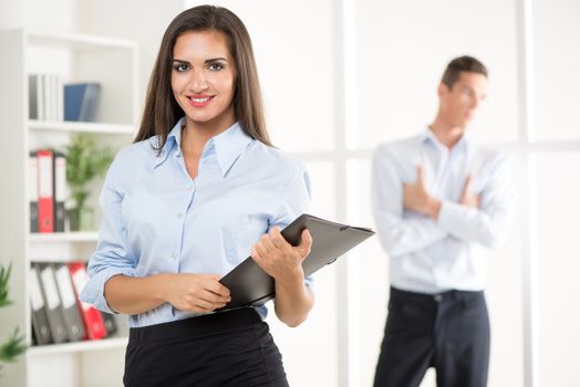 Beautiful young businesswoman standing with document in the office. Colleague standing in background. Selective focus.