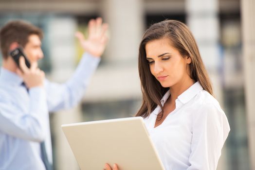 Young business couple standing in front of the building. Business woman looking at laptop while her partner use a mobile phone for a business call. 