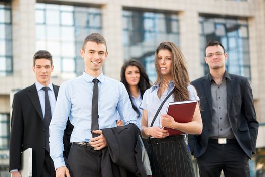 Portrait of group of young successful business people dressed in suits, standing in front of office building, smiling looking at camera.